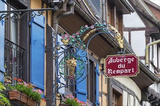 Half-timbered house with blue shutters and flowers. A decorative sign with the inscription 'Auberge