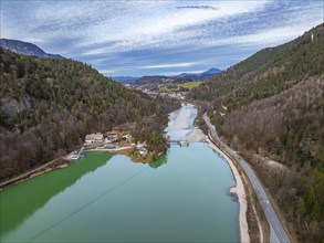 Aerial view of a light blue lake next to a road, surrounded by wooded mountains and some buildings,