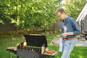 Middle-aged man sprinkles raw steak with oil from bottle while cooking at backyard