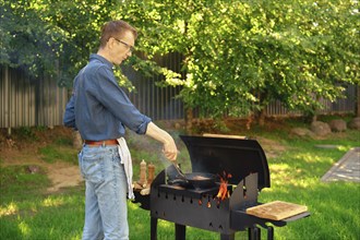 Middle-aged man puts a steak on a frying pan with kitchen tongs while making food at backyard