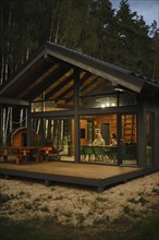 A view from outside at the couple sitting behind dining table in modern forest cabin in the evening