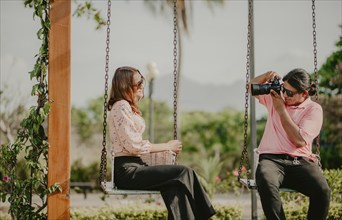 Boyfriend taking a photo of his girlfriend on a swing in a park. Boyfriend photographing girlfriend