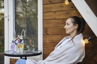 Thoughtful woman sits on terrace wrapped in plaid and enjoys quiet evening