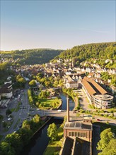 Aerial view of a town with river and bridge, surrounded by forests and hills under a blue summer