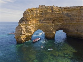 Large rock arch in the blue waters of the Algarve, Portugal, with several boats and tourists in the