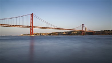 Serene dusk view of a red suspension bridge over calm blue water with reflections, Lissabon, 25 de