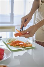 An unrecognizable woman peeling carrots on a green cutting board in a sunlit kitchen. Slices of