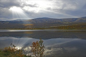 A calm lake with mountains in the background, sunbeams breaking through the clouds, autumn colours