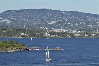 A sailboat sails on a calm sea in front of a hilly landscape with scattered houses and clear blue