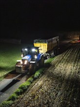 Tractor with trailer driving along a field at night, maize harvest, Dachtel, Black Forest, Germany,