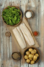 Fresh fish fillets on parchment paper surrounded by bowls of spinach, potatoes, and various spices,