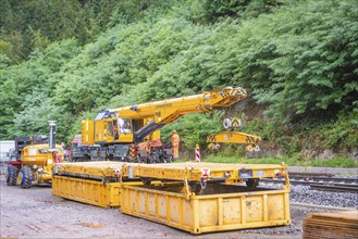 A yellow crane stands next to tracks in the countryside while construction workers work on them,