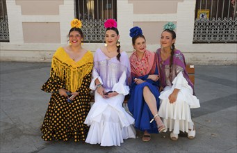 Four woman in colourful flamenco dresses and flowers in their hair sit on a bench on a street in