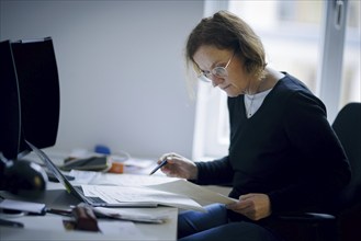 Symbolic photo on the subject of analogue organisation of papers. A woman sits at her desk in the