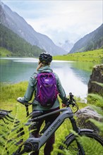 A woman with a helmet and purple rucksack stands in front of a lake, surrounded by mountains, next