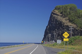 A road along a large cliff with a view of the sea and road signs indicating a cycle path, Gaspesie,