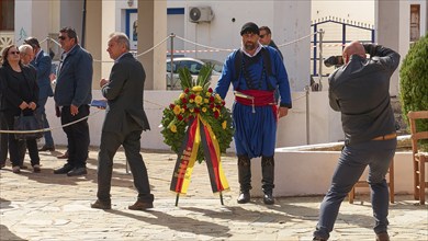 A man in traditional Cretan traditional costume carries a wreath at a ceremony, photographed by a