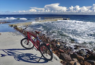 A red bicycle stands on the shore with a view of the rough sea and thick clouds in the blue sky,