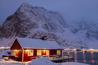 Red wooden house on fjord in front of snowy mountains, illuminated, twilight, winter, Reine,