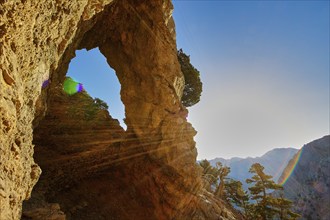 A rocky arch with sunbeams, surrounded by trees and mountains under a clear sky, path to the summit