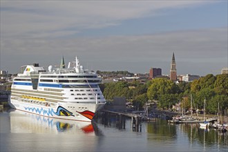 Large white and blue cruise ship in the harbour, with city buildings and a church tower in the