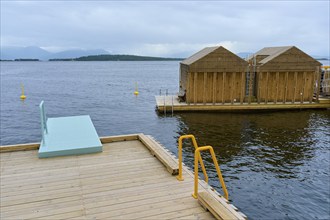 Seaside outdoor pool by the fjord with small wooden sauna huts, overcast sky and calm water, Molde,