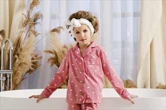 Cute child in a pink pajama and hair band stands next to a modern white bathtub, surrounded by