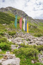 Scene in the mountains with colourful flags, a sign and green vegetation, surrounded by rocks,