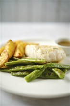 A tender fish fillet alongside fresh green beans and golden roasted potatoes. A small bowl of sauce