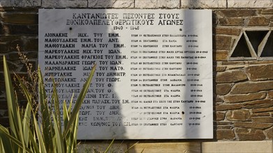 Memorial plaque on stone wall with Greek names, flanked by decorative planting, visit of Federal