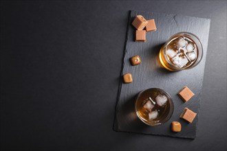 Glass of amber whiskey with ice and caramel candies on a black stone slate board on black