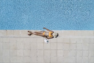 Young woman lying in the sun at the edge of a swimming pool, her face covered by a sun hat,