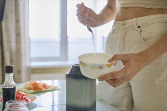 A person adds sugar to a bowl while preparing a meal, surrounded by fresh vegetables and condiments