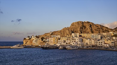 A coastal town with buildings and boats in the harbour under a rocky hill, Pigadia, town and