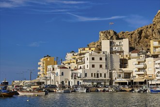 Whitewashed buildings in a coastal town at the harbour with boats and the Greek flag on a hill in