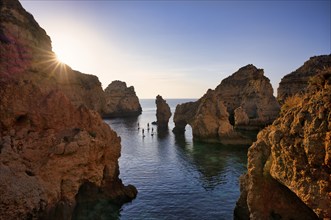 Stand Up Paddlers on their boards, colourful cliffs and sunrise, rocks in the sea, Ponta da