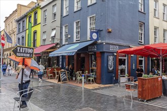 Colourful street scene with cafés and people walking under umbrellas, Cork