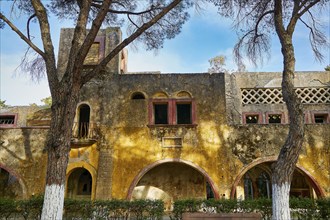 Ruined building with yellow façade and arches flanked by tall trees with blue sky, Italian