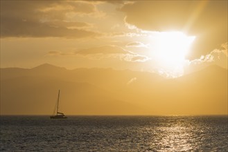 A sailboat on a calm sea at sunset with a golden sky and mountains in the background, Agia Kyriaki,