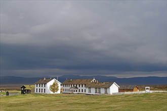 Landscape with white buildings in an open field against a cloudy sky and mountains, Foksummyra,