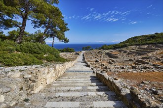 Stone steps lead through ruins with views of the nearby sea and surrounding vegetation under a blue