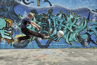 A ten-year-old boy plays with his football in front of a graffiti wall, Germany, Europe