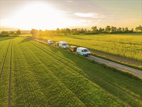 Cars driving along a rural road next to green fields at sunset, fibreglass installation, Calw