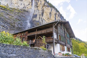 Rustic wooden house in front of an impressive waterfall, surrounded by lush greenery and autumn