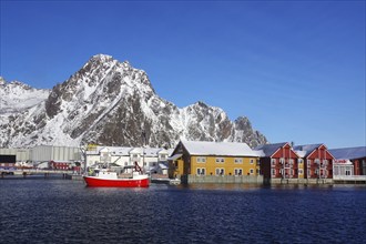 Colourful houses and a red fishing boat in the harbour in front of snow-covered mountains under a