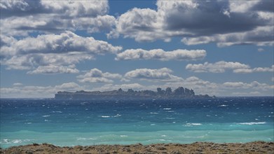 View of an island in the sea under a cloudy sky with blue water and coastal vegetation, islet of