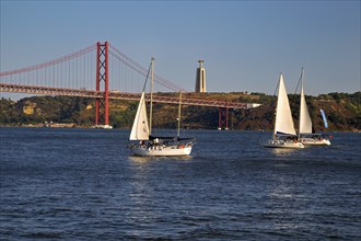 Two sailboats on calm waters with a red suspension bridge in the background under a blue sky,