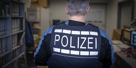 Police officer in office, back view, surrounded by files and computers, symbolic image search,