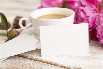 White business card with pink peony flowers and cup of coffee on white wooden background. side