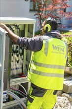 A worker in a safety waistcoat is busy with fibre optic assembly, surrounded by a garden with a red